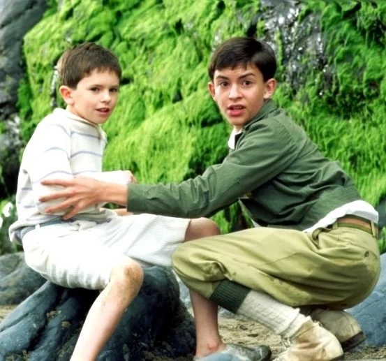 Dos niños en una escena de película, uno de ellos se sienta en una roca y el otro parece estar sosteniéndolo, con un fondo natural de vegetación verde.